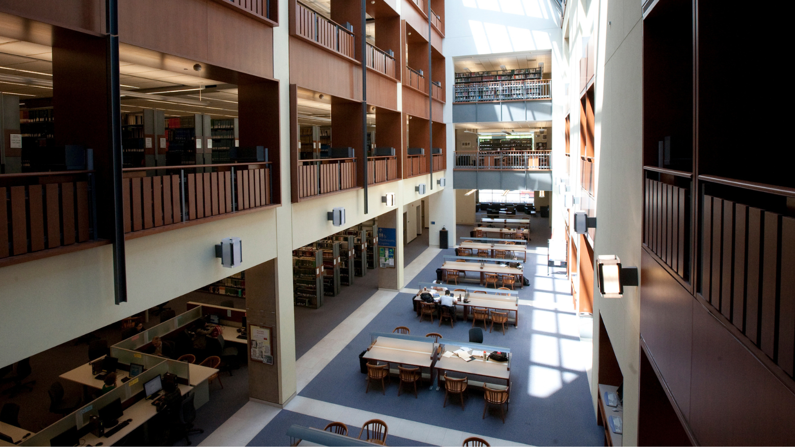 a view of Stauffer library at Queen's main floor, taken from the second floor balcony. Sunlight is streaming through an out of shot skylight and onto the wooden tables and blue floors