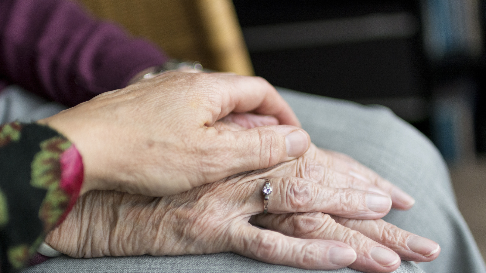 a close up shot of an elderly adult's hand with a diamond ring on it, while another elderly person's hand lovingly rests on top of it in a supportive gesture.