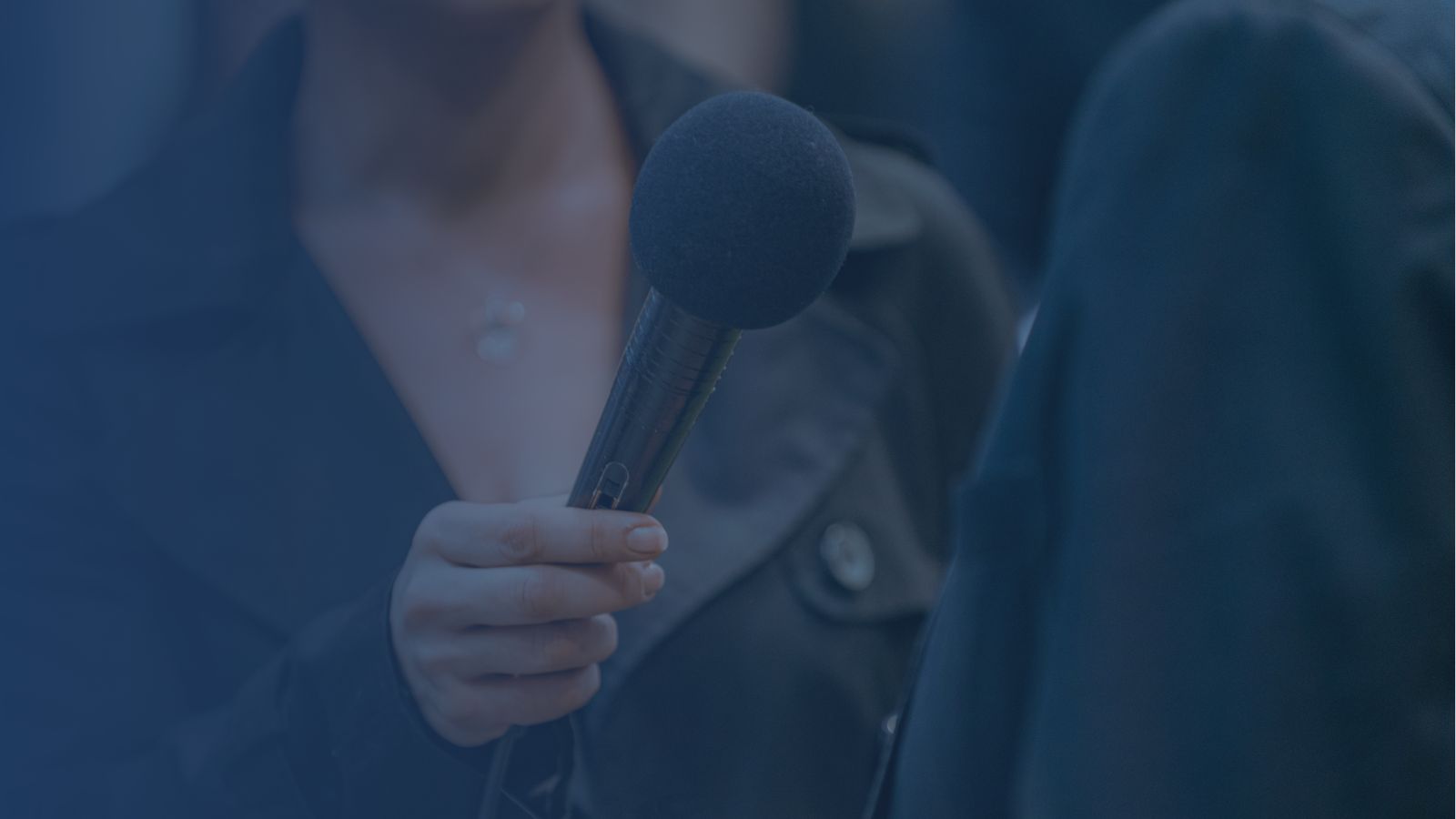 female reporter wearing a jacket and holding up a microphone to an interviewee, whose back is to the camera. Only their torsos are in the frame