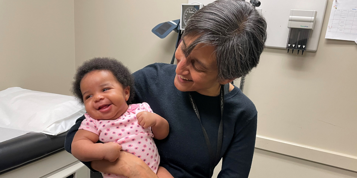 healthcare professional Dr. Rupa Patel holding a smiling baby in a medical examination room.