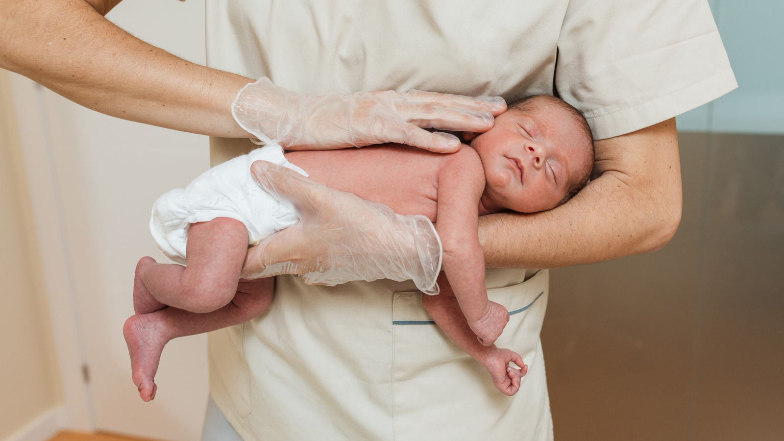 a newborn lying on their stomach, being held by a healthcare provider in ivory scrubs and wearing clear gloves.