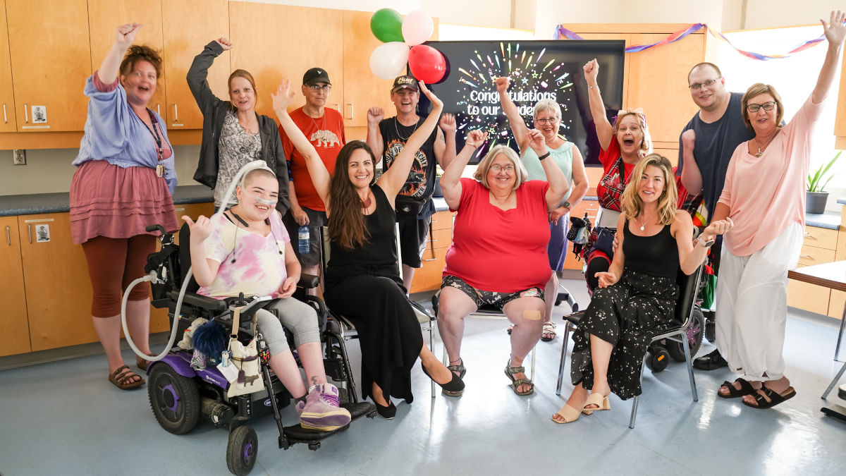 Class photo of a group of diverse individuals, smiling and cheering. Some are throwing their hands up in celebration above their heads. Recovery College students wrapping up their semester at a celebration party.