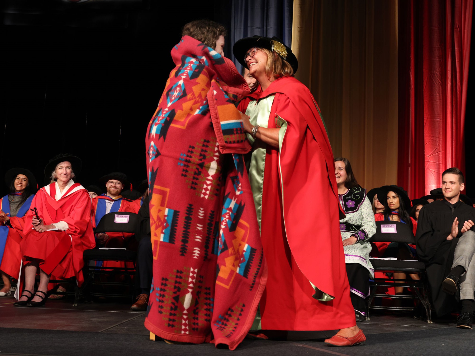 A graduating indigenous undergraduate nursing student being wrapped in a ceremonial blanket by the director of the school of nursing, who is wearing red and gold academic regalia. They are both smiling as they look at each other.