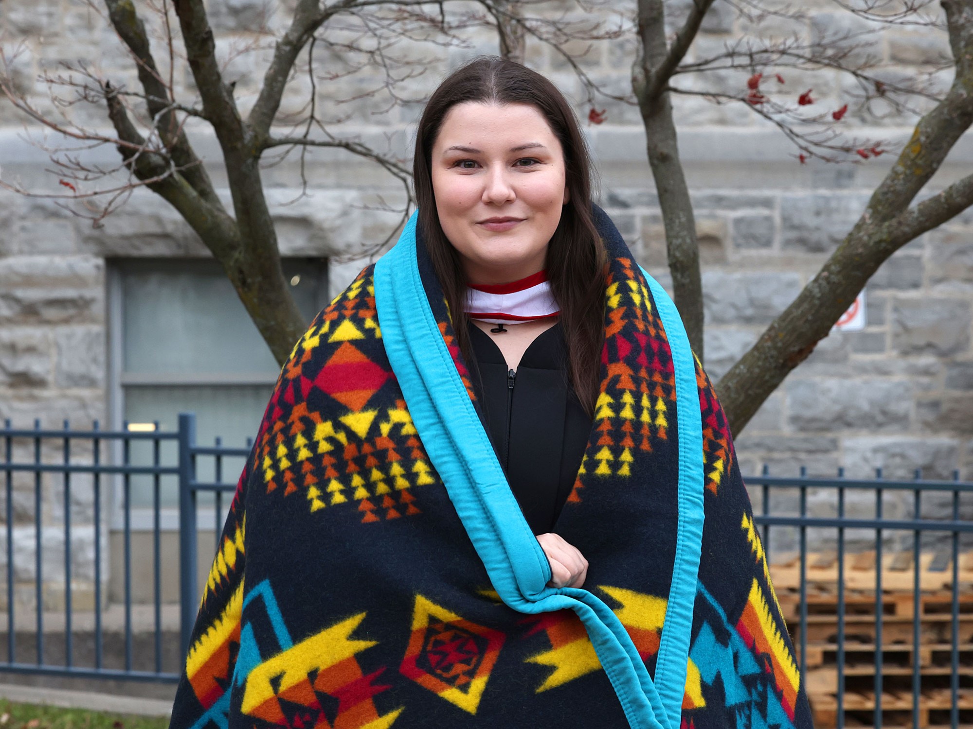 An indigenous undergraduate nursing student wrapped in a ceremonial blanket and wearing convocation robes, standing together in front of a limestone building and smiling at the camera