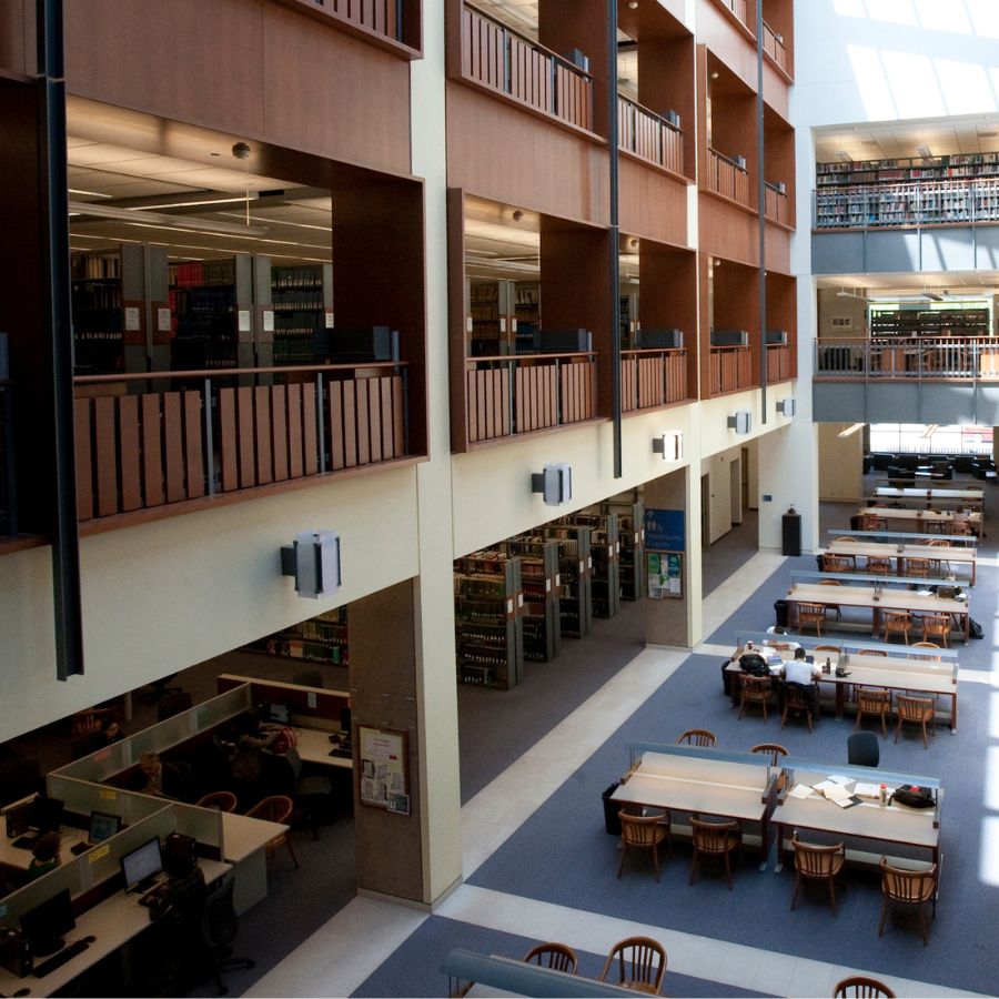 a view of Stauffer library at Queen's main floor, taken from the second floor balcony. Sunlight is streaming through an out of shot skylight and onto the wooden tables and blue floors