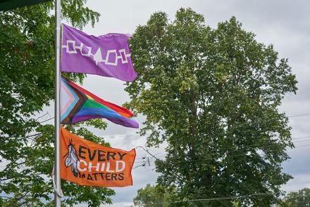 Photo of the Iroquois Confederacy Flag, a Pride Flag, and an Every Child Matters Flag on a flag pole.
