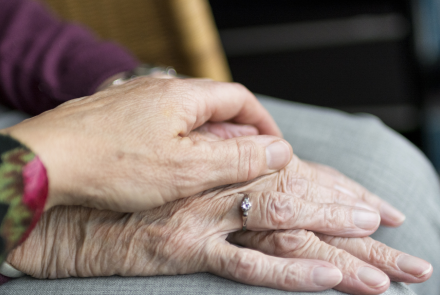 a close up shot of an elderly adult's hand with a diamond ring on it, while another elderly person's hand lovingly rests on top of it in a supportive gesture.