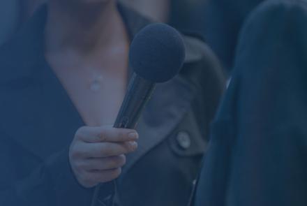 female reporter wearing a jacket and holding up a microphone to an interviewee, whose back is to the camera. Only their torsos are in the frame