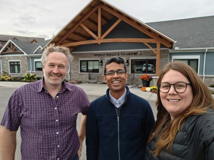 Drs. Craig Goldie, Aynharan Sinnarajah and Katie Goldie smiling side by side in a group photo in front of the exterior stone entryway if Oak Ridges Hospice in Port Perry during a site visit in Fall 2024