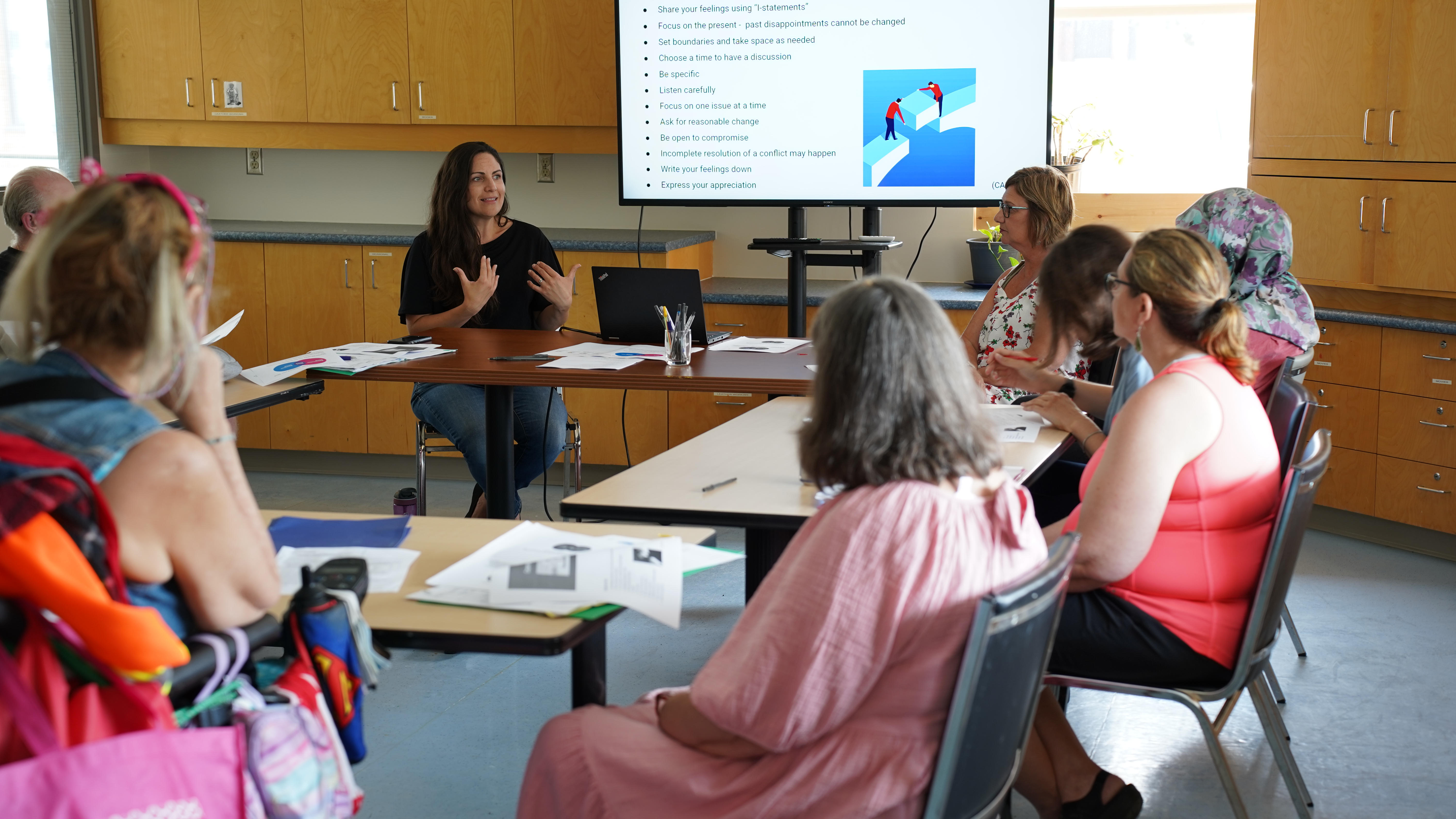 Ellie Lambert sitting at a round table of desks talking animatedly with a group of students who are listening to her speak.